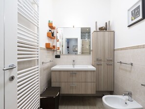 A modern bathroom with a white sink, mirror, and cabinet, a bidet, and a small trash bin.