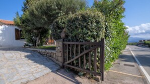 A stone pathway leading to a wooden gate, surrounded by lush greenery and a white building with a tiled roof. - Beautiful apartment in villa for 5 guests with WIFI, A/C, TV, patio and pets allowed (Arzachena)