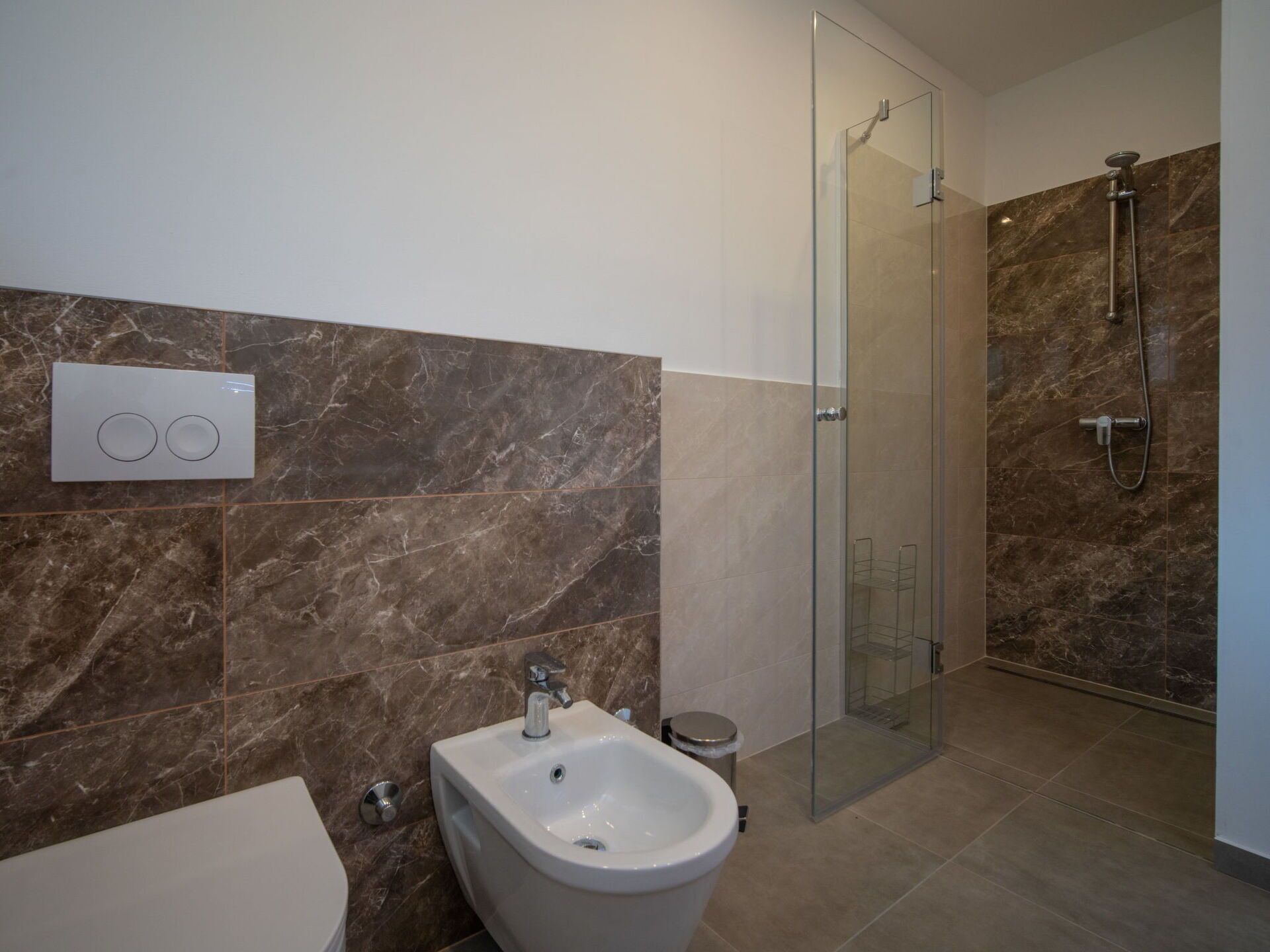 Een badkamer met een bidet, een douche met glazen wanden en een wand betegeld met marmer.