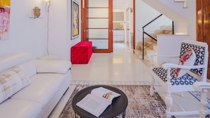 A modern living room with a white sofa, a red ottoman, a patterned armchair, and a staircase leading to an upper floor.