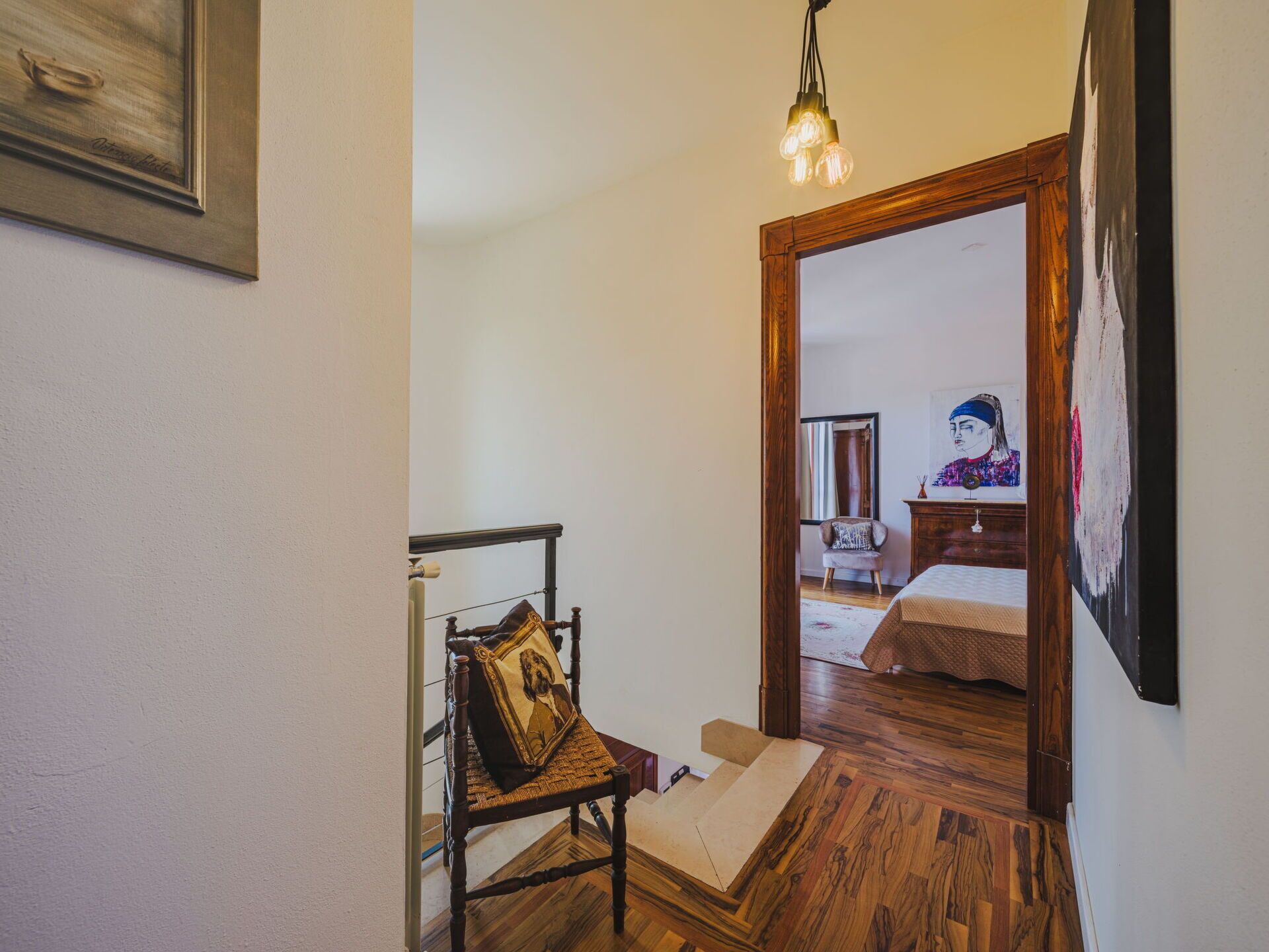 A hallway with a wooden floor, a chair with a cushion, a hanging light fixture, and a mirror reflecting a room with a bed and a painting.