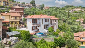 A multi-story house with a pool, surrounded by lush greenery and other residential buildings.