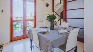 A dining area with a table set for two, a vase with flowers, and a view of a building through a large window.
