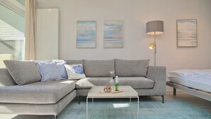 A modern living room with a grey sectional sofa, a white coffee table, and a floor lamp. There are three paintings on the wall and a bed in the background.