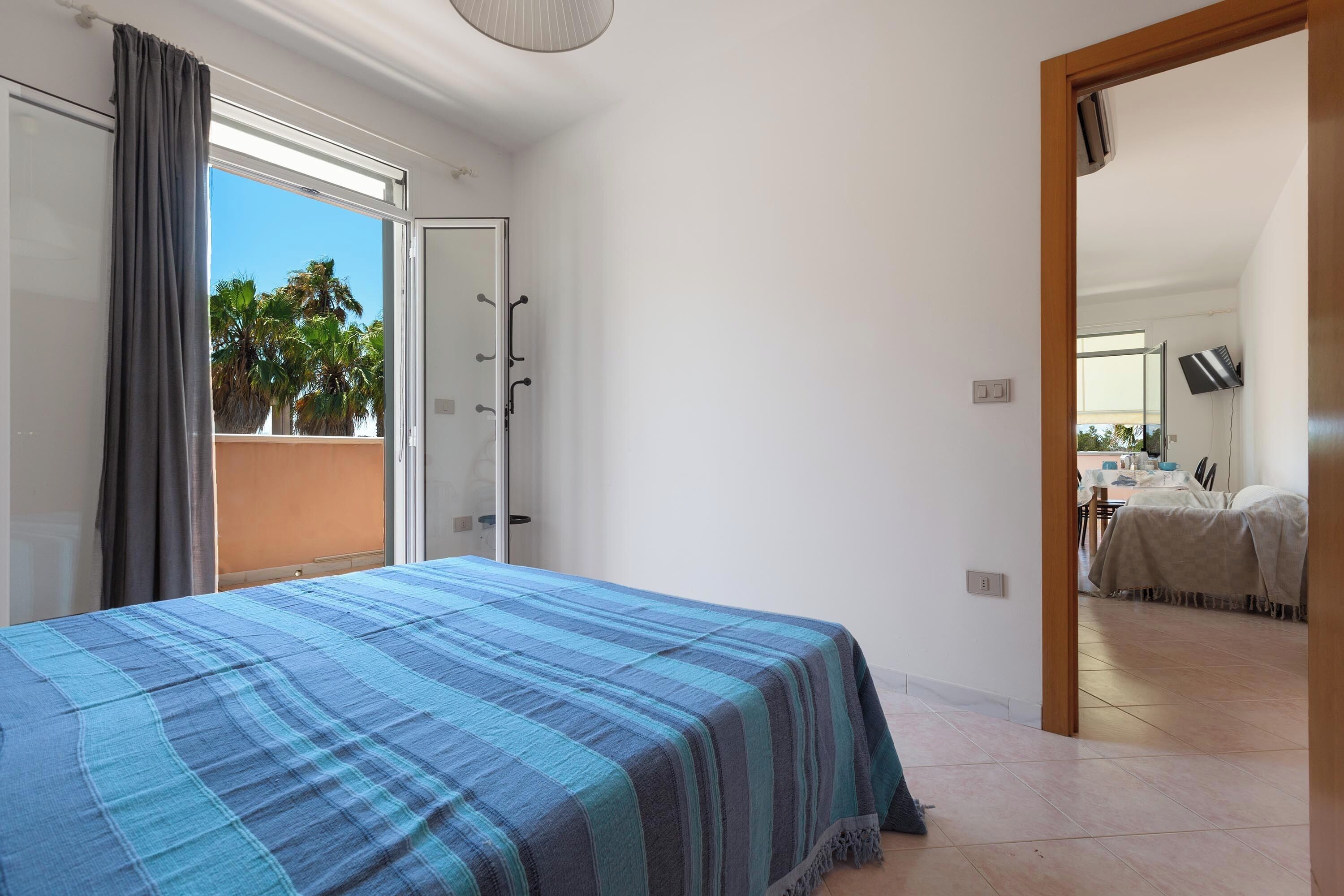 A bedroom with a bed, a sliding glass door to a balcony, and a view of palm trees.