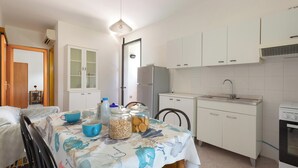 A compact kitchen with white cabinets, a sink, and a stove. There is a dining table with a tablecloth and various kitchen items on it. - 4128 Villino Giovanna by Barbarhouse (Melendugno)