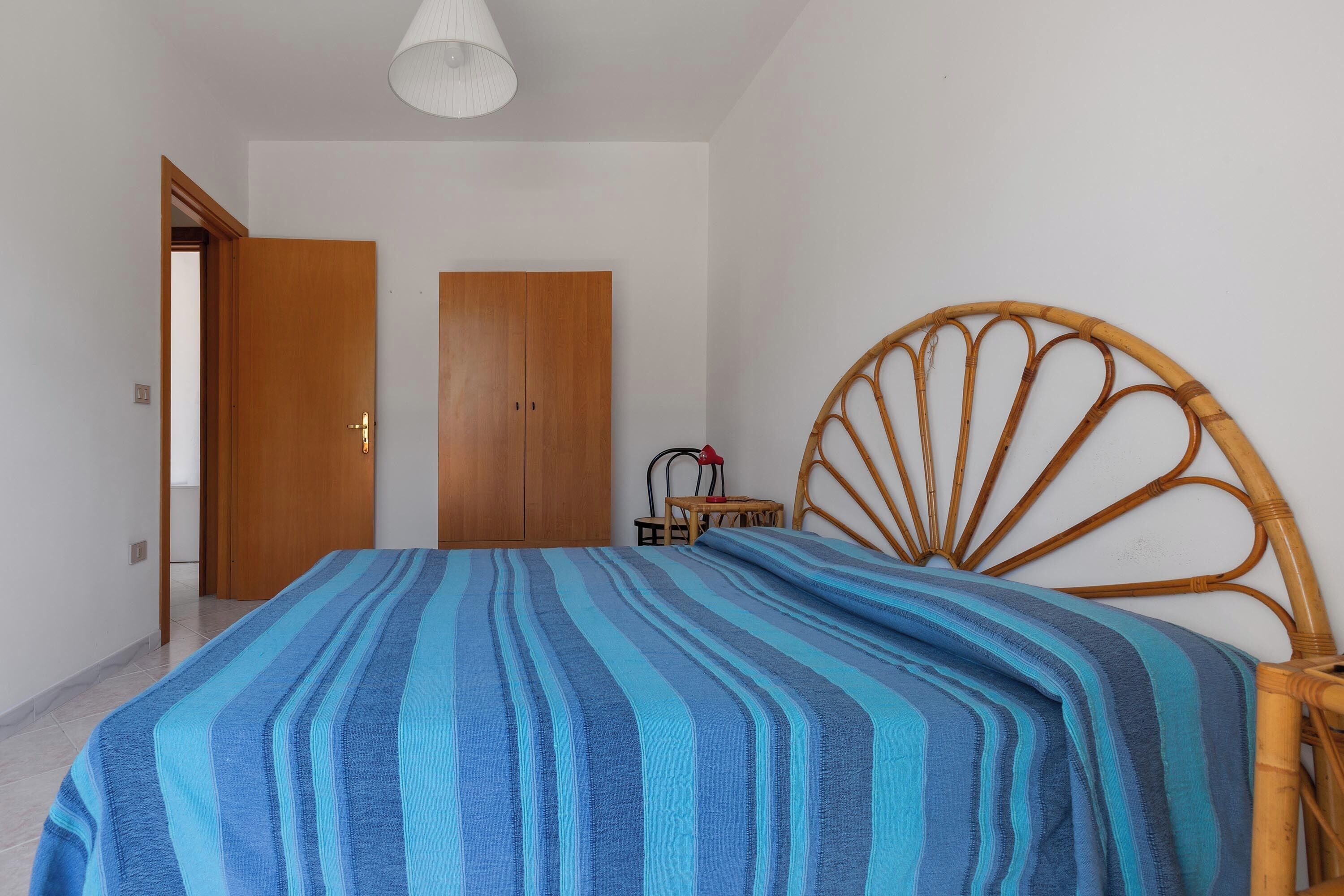 A bedroom with a bed, a wooden wardrobe, a wicker chair, and a white ceiling light.