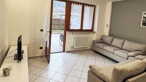 A living room with a sofa, a TV, a radiator, and a window with curtains. - Cosy apartment for 4 people with WIFI, TV, patio and pets allowed (Pieve Di Ledro)