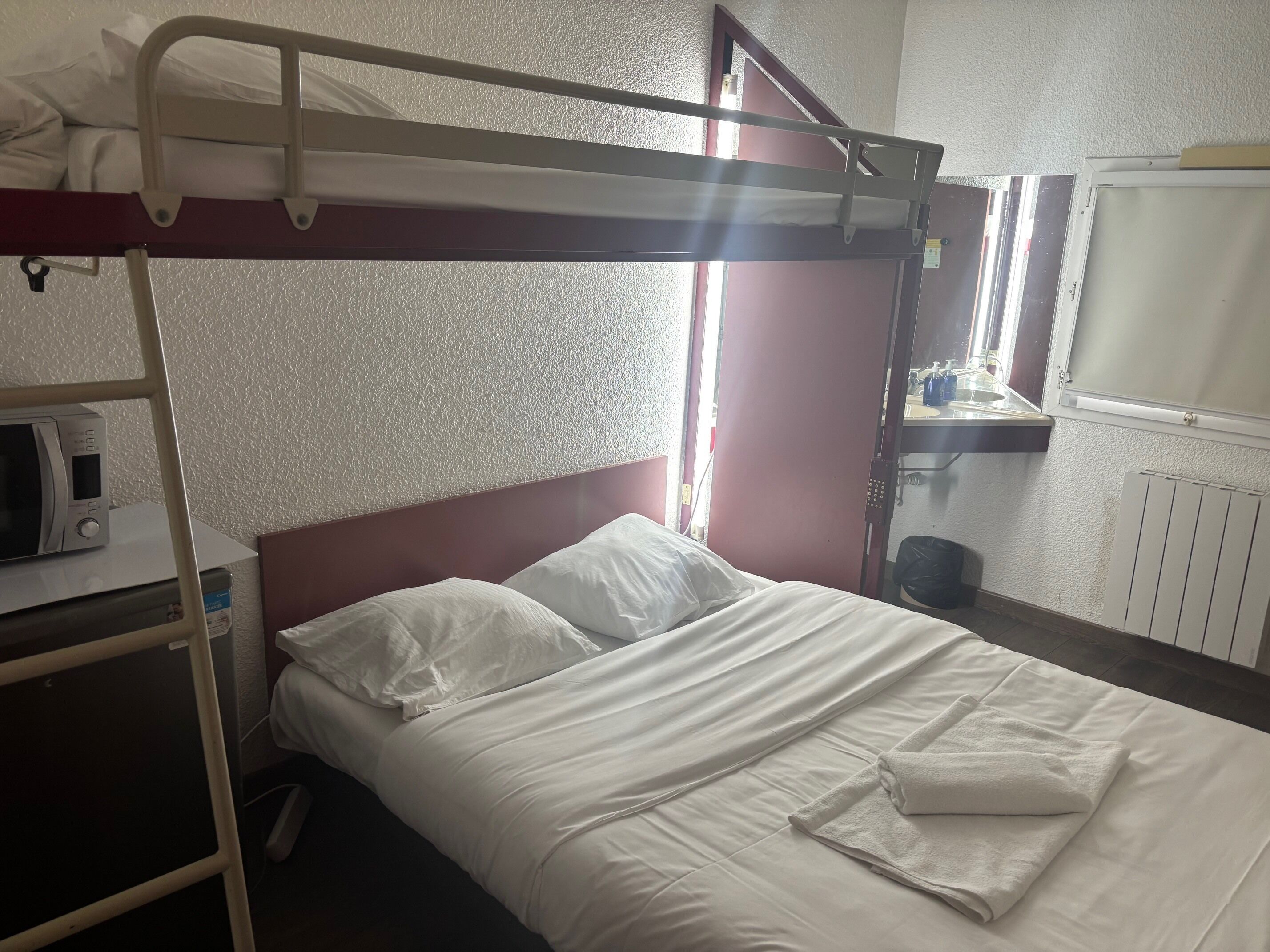 Una cama litera con ropa de cama blanca, un microondas sobre una mesita y una ventana con cortinas.