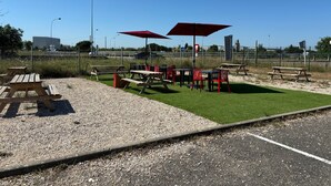 An outdoor seating area with picnic tables and red umbrellas, surrounded by a fence and a gravel path.