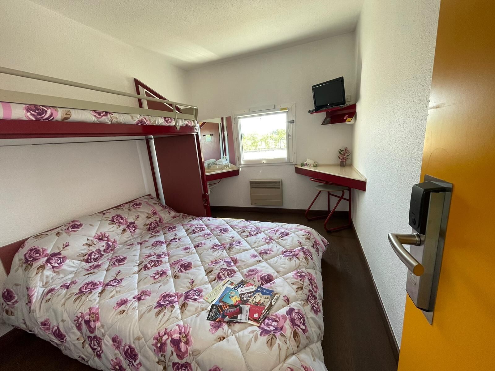 Una cama litera con una colcha estampada, un escritorio con monitor de computadora y una ventana con vista a follaje.