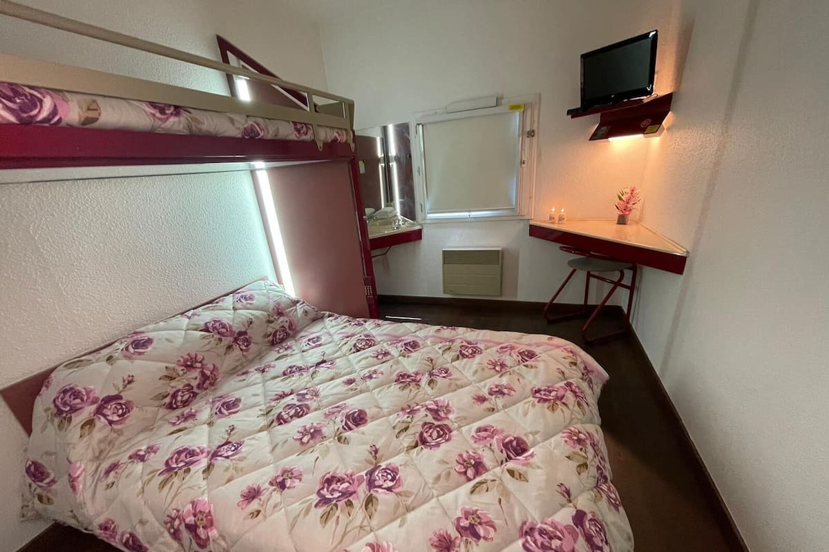 A bedroom with a bunk bed, a desk, a chair, a TV, and a window.