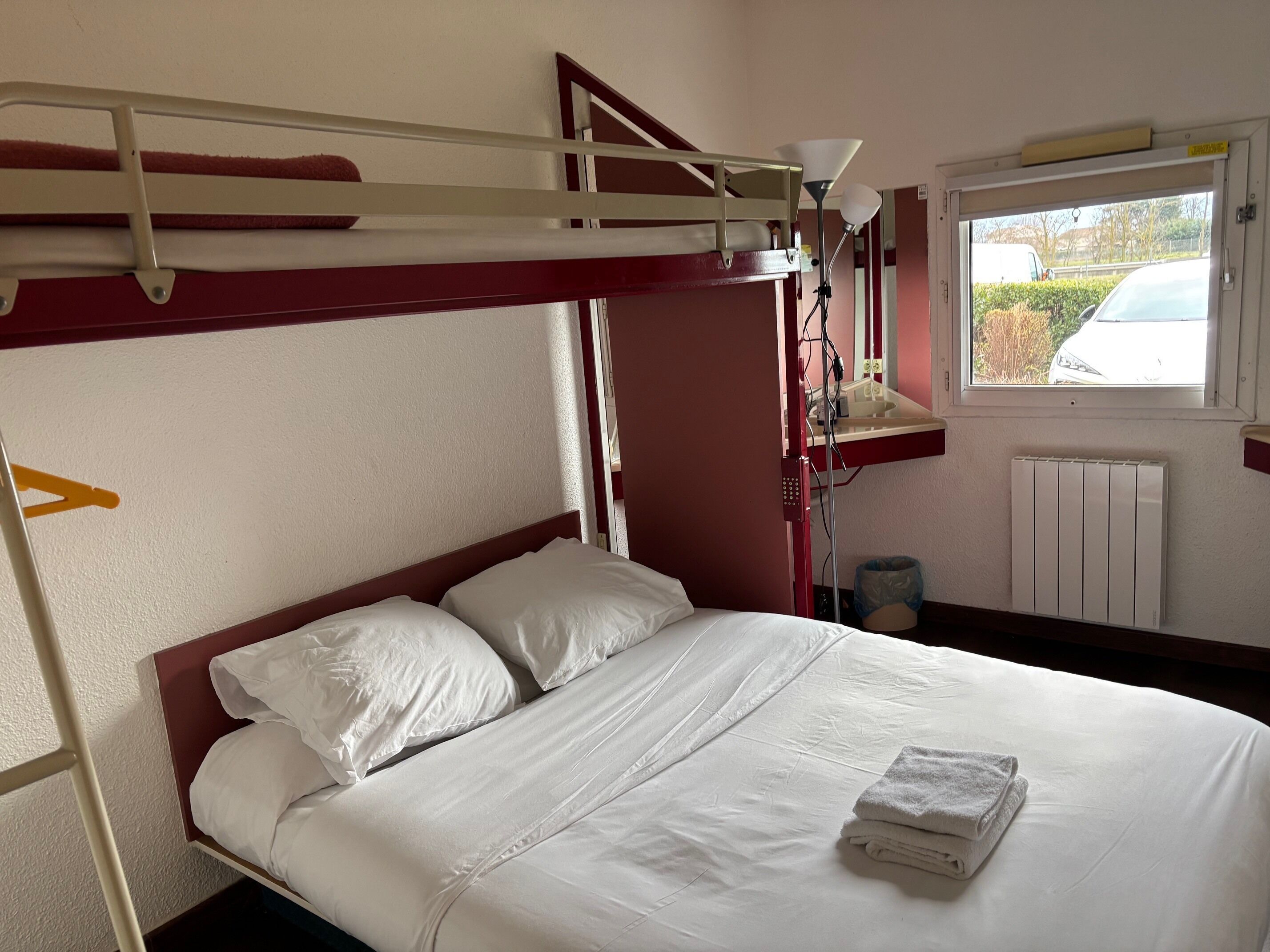 A bunk bed with white bedding, a bedside lamp, and a window with a view of a car and trees.