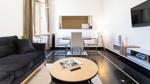 Ein modernes Hotelzimmer mit einer schwarzen Couch, einem runden Couchtisch, einem Schreibtisch mit Stuhl, einem an der Wand montierten Fernseher und einer kleinen Pflanze.