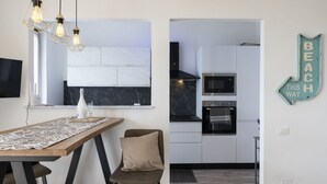 Een moderne keuken met witte kasten, een zwart aanrecht en een ingebouwde oven. Er is een eethoek met een houten tafel en stoelen, en een wandgemonteerde airconditioning.