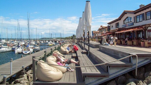 Un muelle de madera con zonas de estar, un puerto deportivo con barcos y edificios con terrazas.
