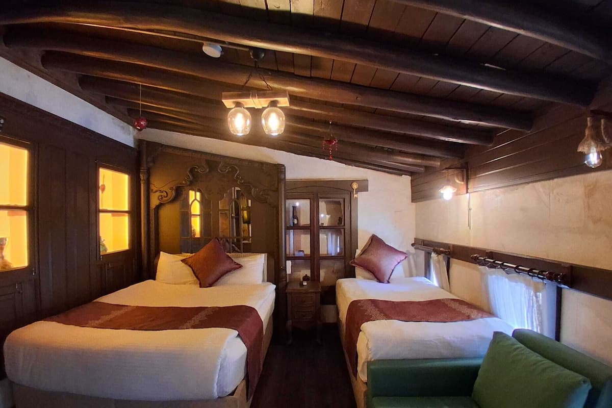 Quarto com duas camas, um sofá verde, vigas de madeira no teto e uma pequena mesa com abajur.