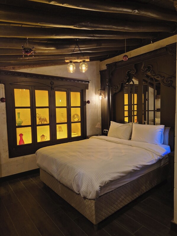 Ein Schlafzimmer mit einem großen Bett, einem Fenster mit dekorativen Bleiglasverzierungen und einer Decke mit freiliegenden Balken und Pendelleuchten.