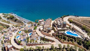 An aerial view of a coastal residential area with multiple swimming pools, a curved road, and a beach in the background.