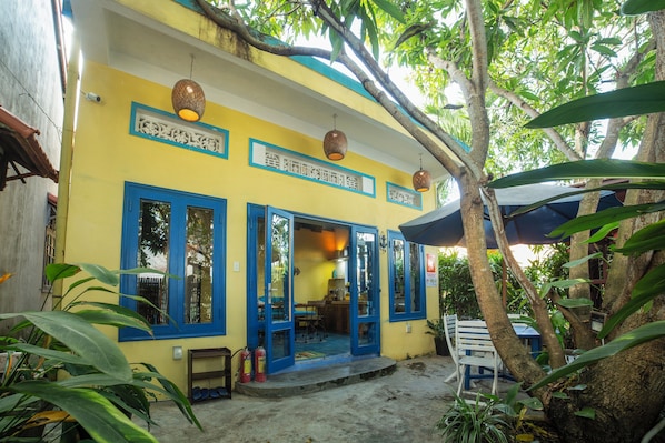 A yellow building with blue-framed windows and doors, a covered outdoor seating area, and a tree in the foreground. - Loft House 2BR Seaside An Bang Beach (Da Nang)