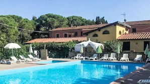 A swimming pool with lounge chairs and umbrellas, surrounded by buildings with red-tiled roofs and greenery in the background.