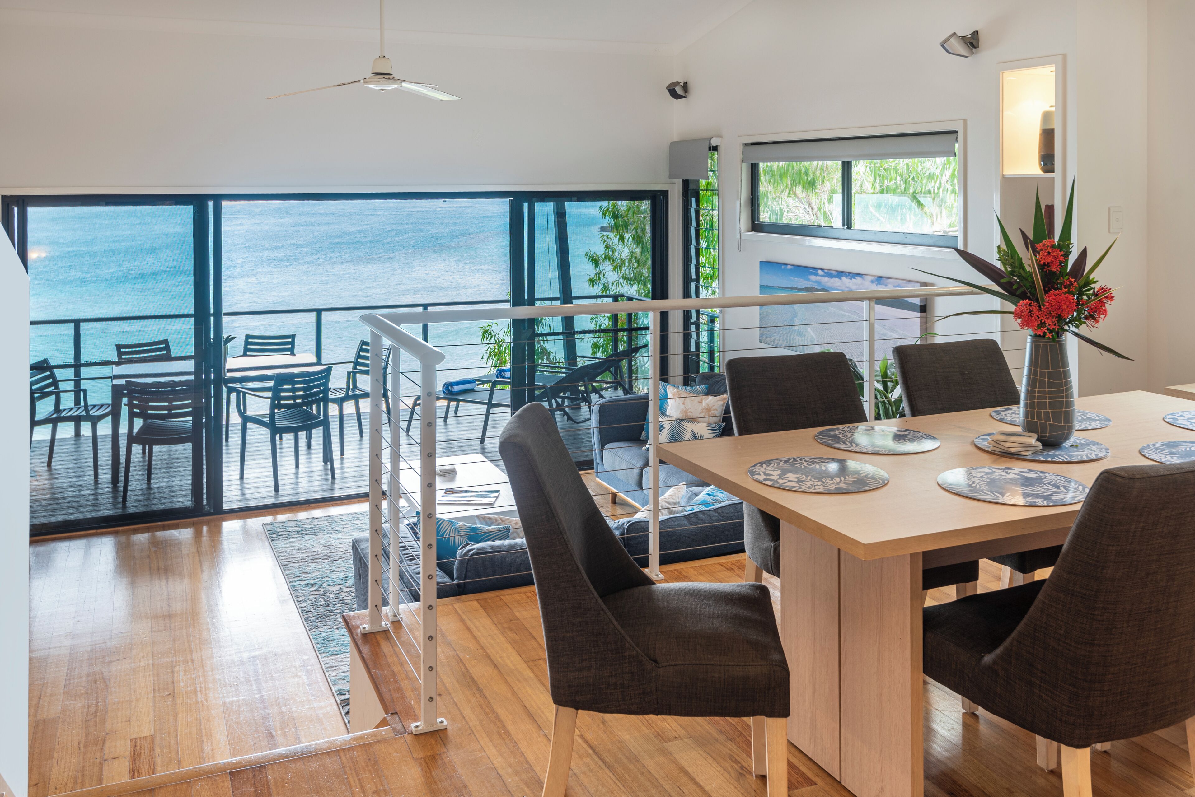 Un coin repas avec vue sur la mer, un balcon avec des chaises et une table dressée avec des assiettes et un vase de fleurs.