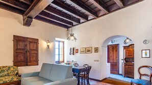 Ein gemütliches Wohnzimmer mit Sofa, Esstisch und Stühlen. Es gibt eine Holzbalkendecke und ein großes Fenster.