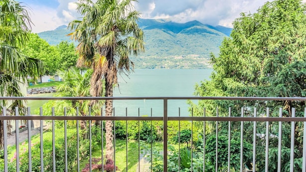 A balcony with a metal railing overlooking a lake, surrounded by lush greenery and mountains in the background.