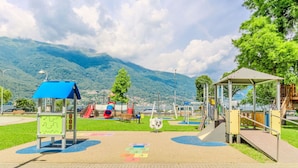 A playground with slides and climbing structures, surrounded by a paved area and greenery, with mountains in the background.