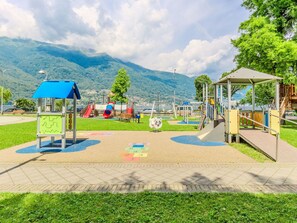 Explore the playground area with slides, climbing frames, and a playhouse, surrounded by a paved path and scenic mountain views.
