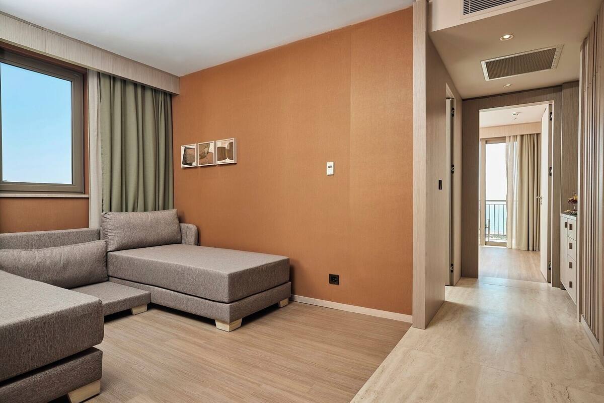 A modern hotel room with a sectional sofa, a large window with curtains, a wall-mounted artwork, and a hallway leading to a balcony.