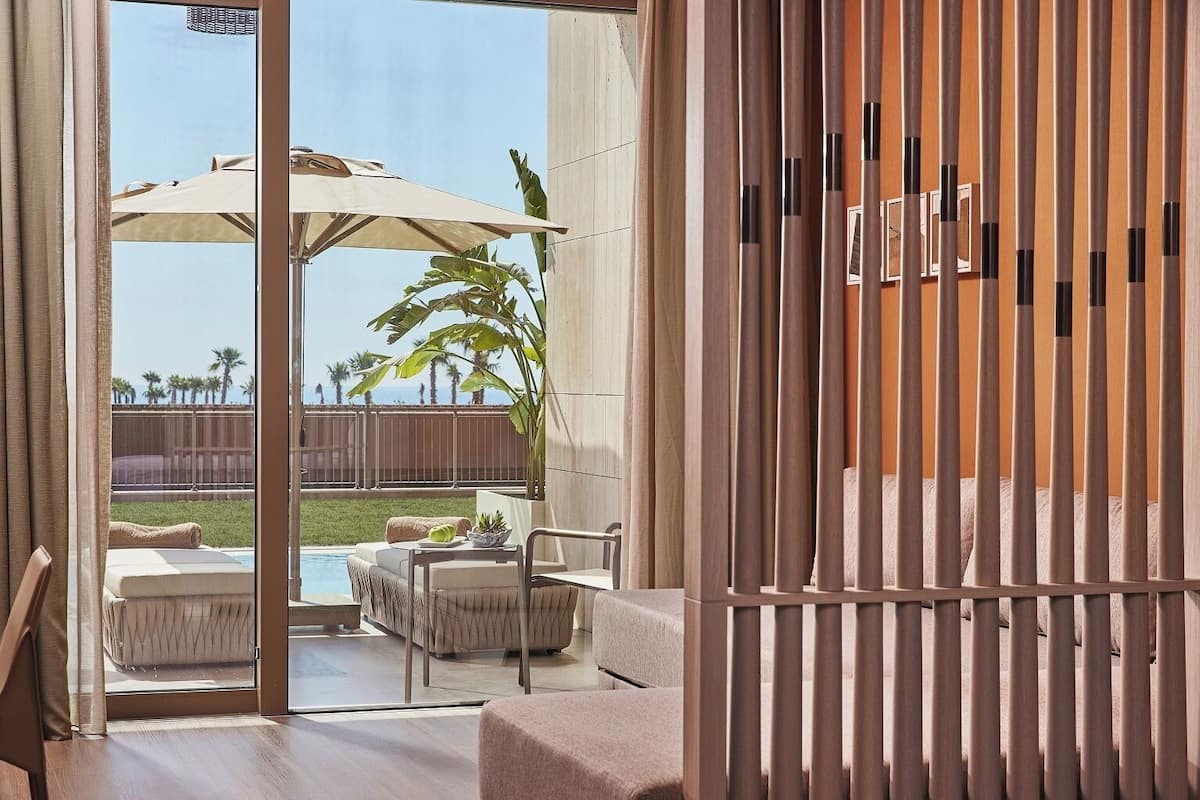 A modern outdoor patio with a pool, seating area, and a view of palm trees and a fence.