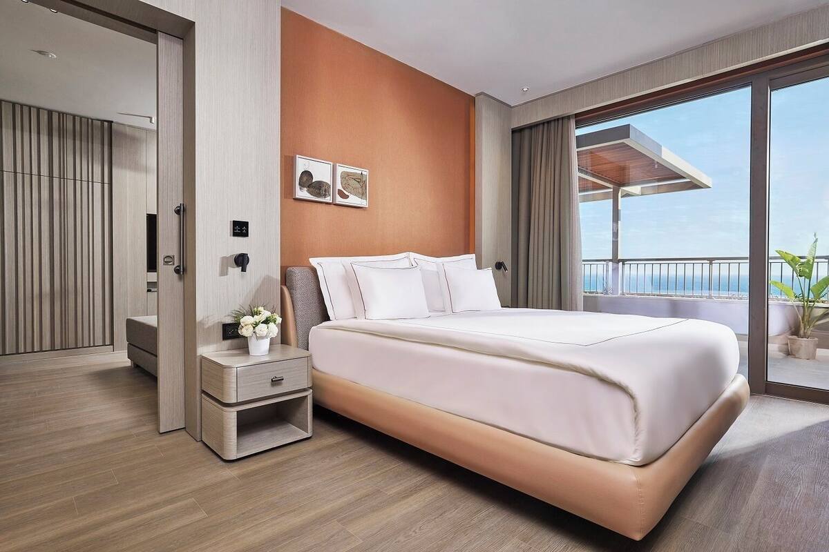 A modern hotel room with a large bed, a nightstand, a small table with a vase of flowers, and a view of the ocean through large windows.