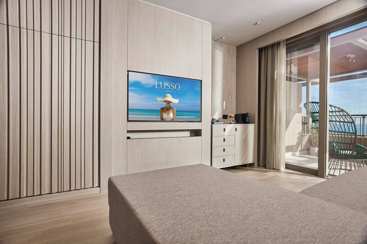 A modern room with a large TV displaying 'LUSSO' and a beach scene, a sofa, a small cabinet, and a sliding glass door leading to a balcony with outdoor furniture.