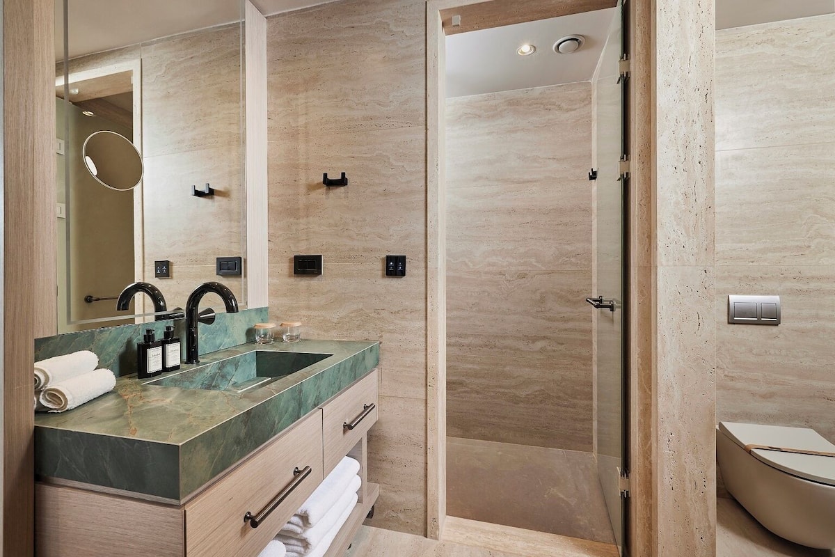 A modern bathroom with a dual sink vanity, a large mirror, a walk-in shower, and a toilet.
