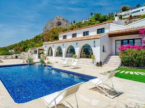 A poolside area with a clear blue pool, white lounge chairs, and a white building with arched windows and a tiled roof. - Beautiful private villa for 8 guests with WIFI, A/C, private pool, TV and patio (Xàbia)
