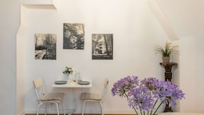 Ein gemütlicher Raum mit Esstisch für zwei Personen, einem kleinen Beistelltisch mit einer Vase voller lila Blumen und drei schwarz-weißen Waldmotiven an der Wand.