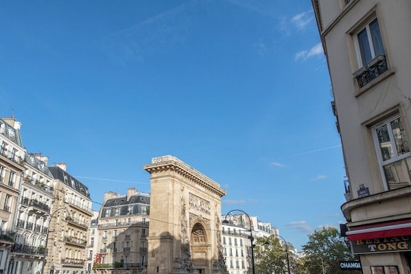 A historic stone monument in an urban setting with classical architecture, surrounded by buildings with balconies and storefronts.