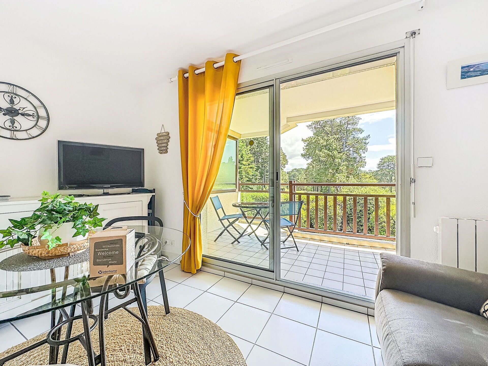 Ein Zimmer mit Fernseher, Glastisch, Stühlen, einer Couch, einem gelben Vorhang und einer Glasschiebetür, die auf einen Außenbereich mit einem Balkon führt.