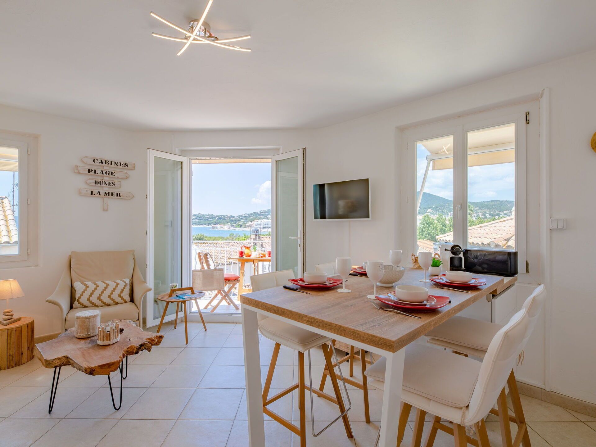 A dining area with a wooden table, white chairs, and a view of the sea through open doors.