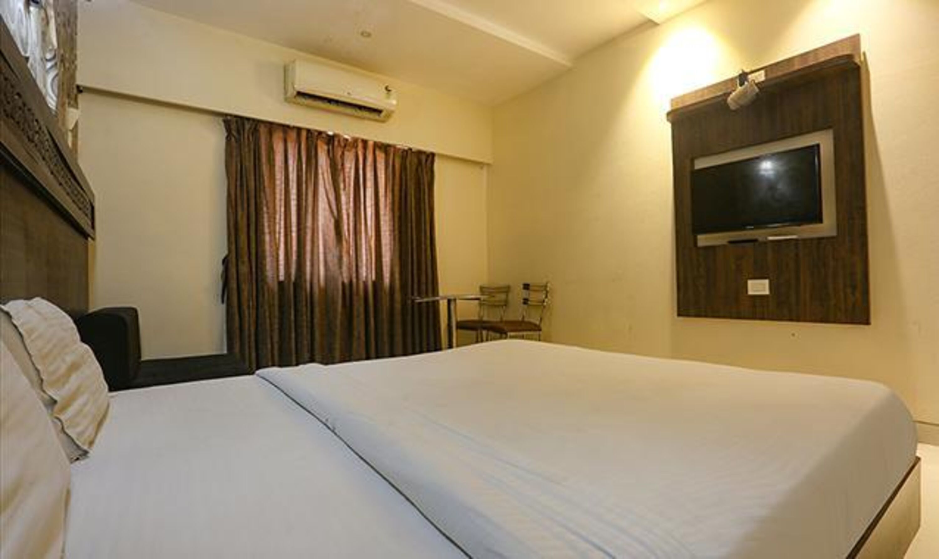A hotel room with a large bed, a TV mounted on the wall, a desk with two chairs, and a window with curtains.