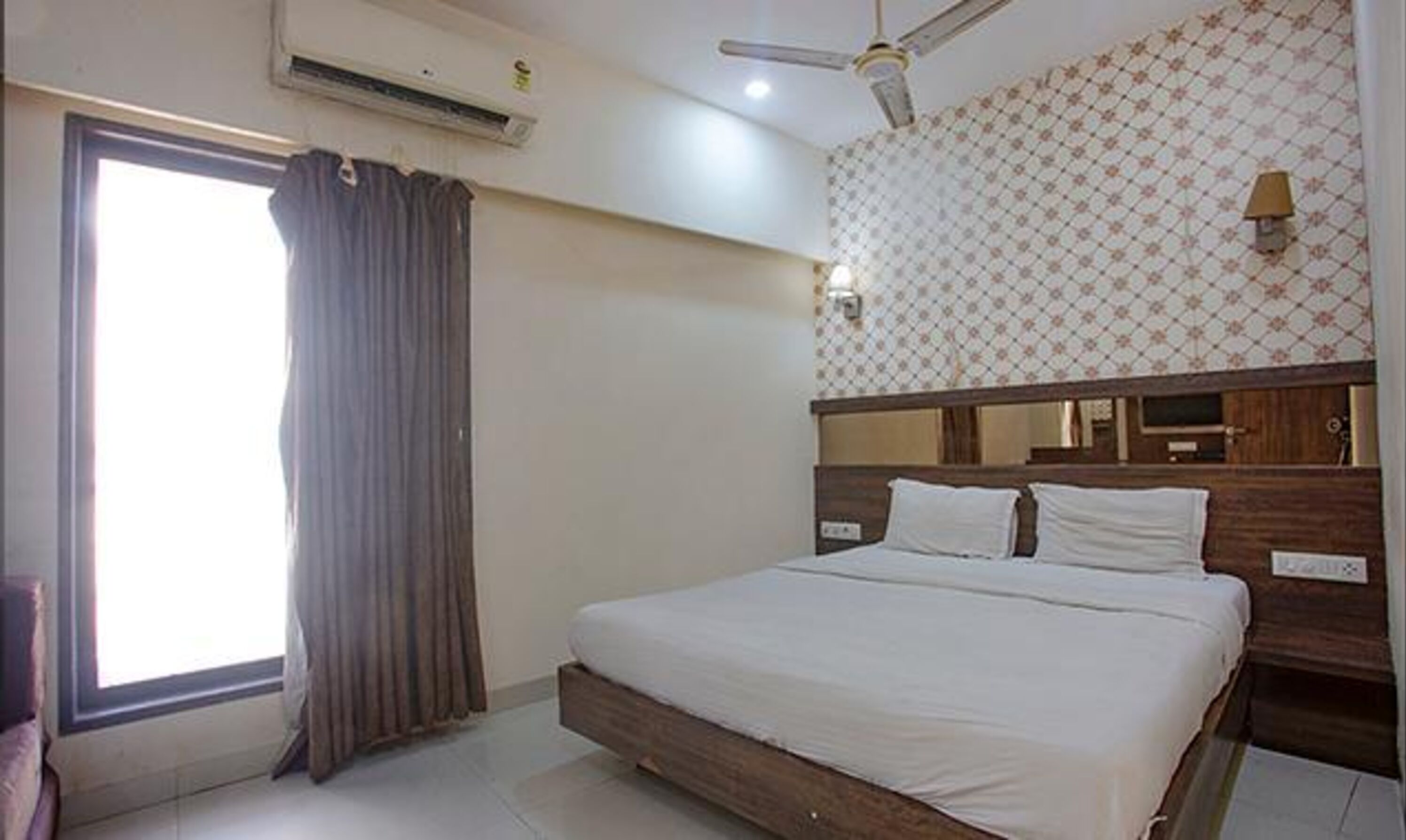 A hotel room with a large bed, a ceiling fan, an air conditioning unit, a window with curtains, and a patterned wall.