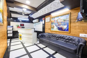 Un vestíbulo de hotel con un sofá de cuero negro, un mostrador de recepción, un cuadro de un puerto y un reloj digital.