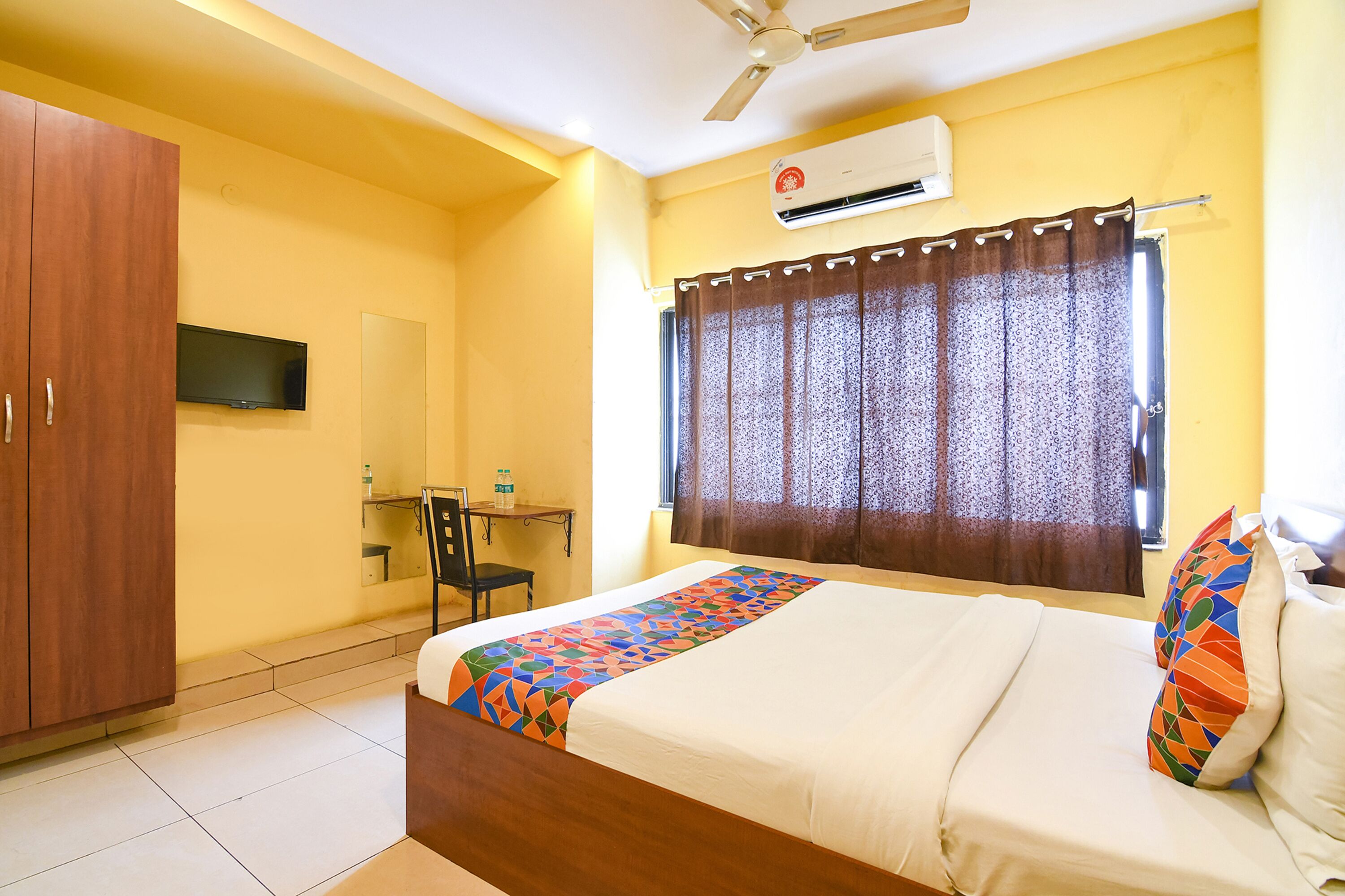 A hotel room with a bed, a wardrobe, a desk with a chair, a television, and an air conditioning unit.