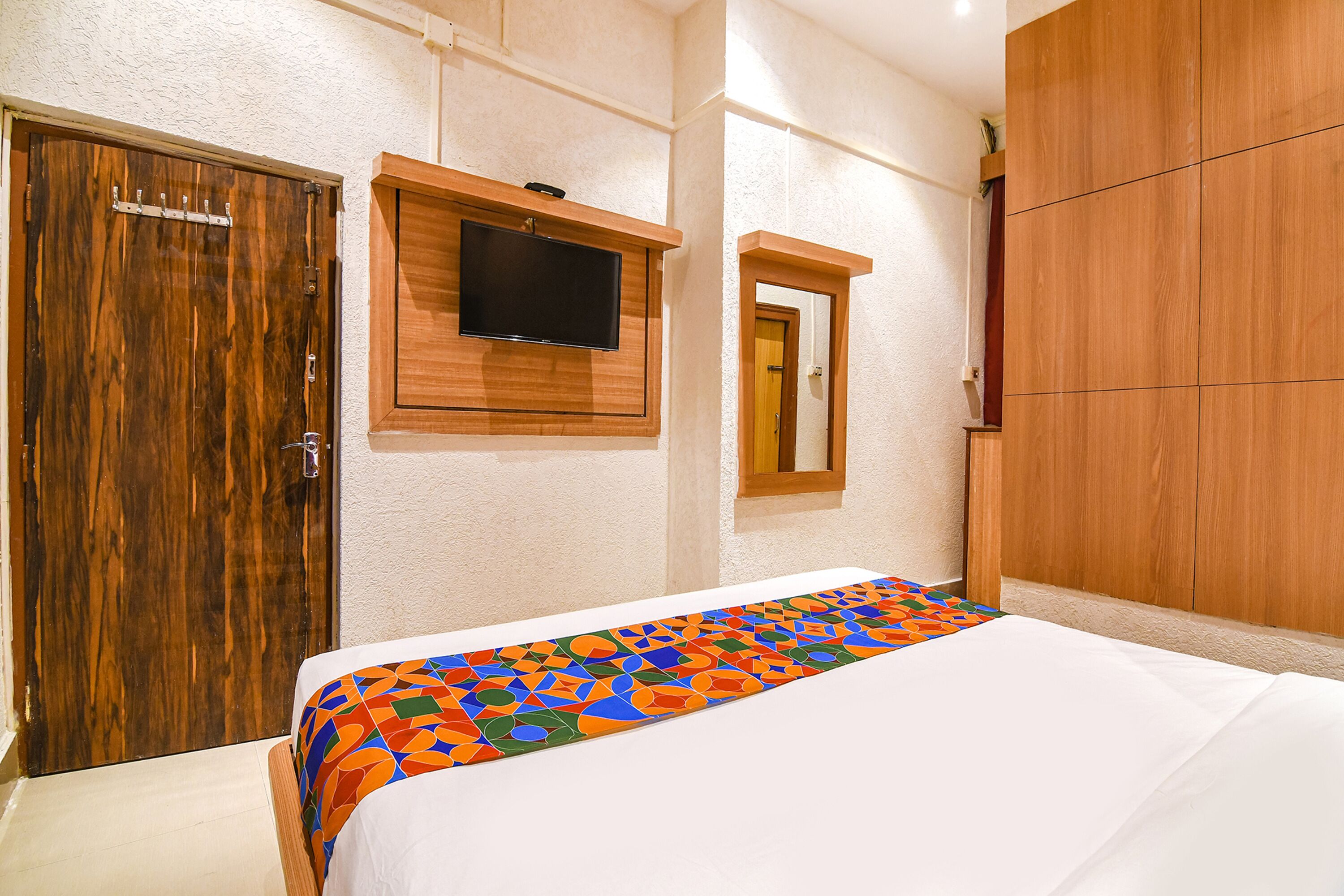 A bedroom with a bed, a wooden wardrobe, a television mounted on the wall, and a small window.