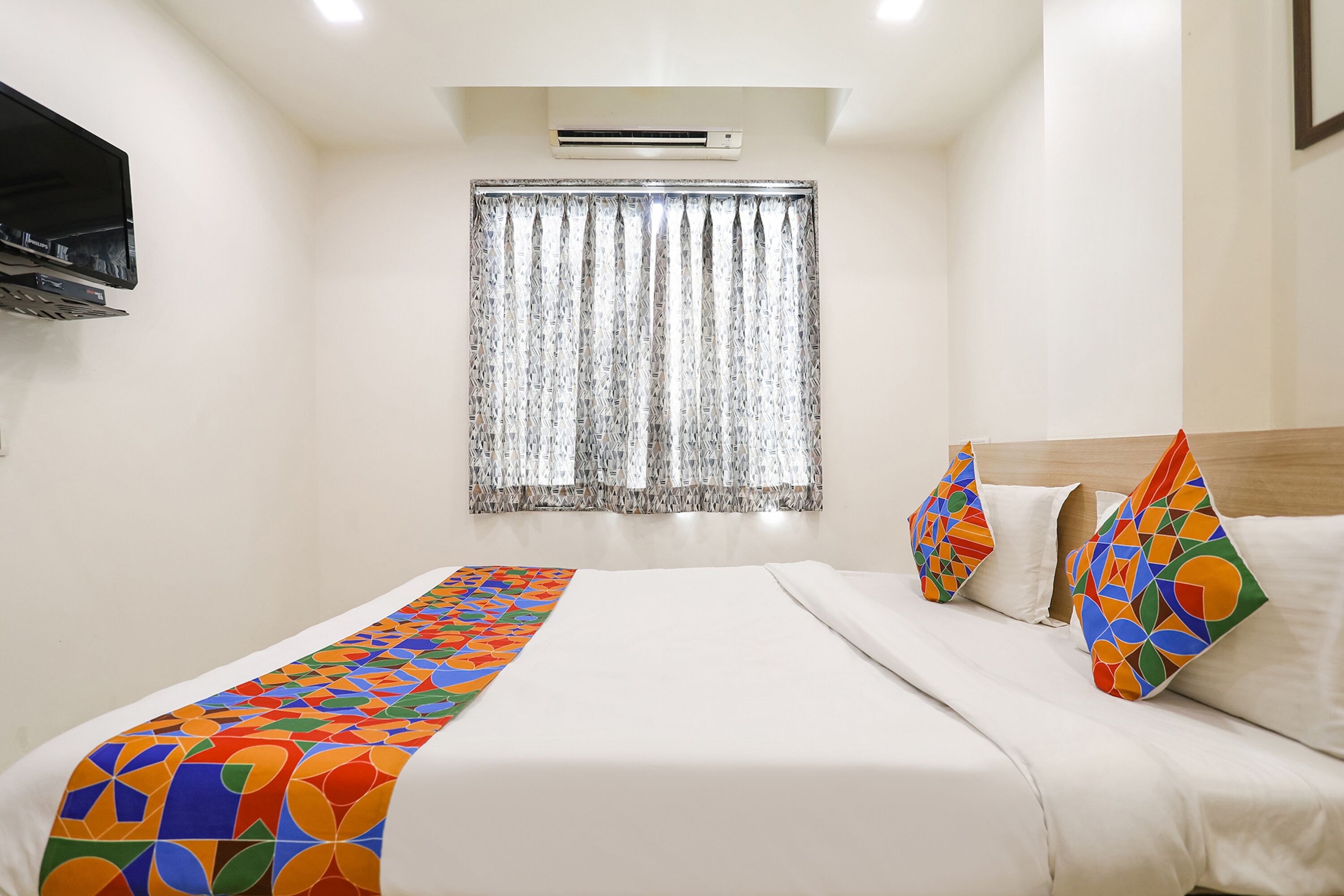 Habitación de hotel con cama, televisor montado en la pared, ventana con cortinas y colcha con estampado geométrico de colores.