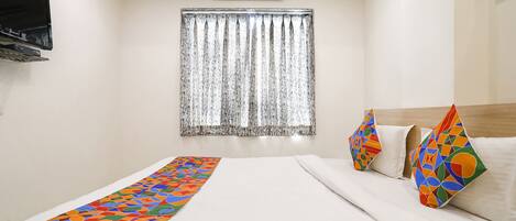 Habitación de hotel con cama, televisor montado en la pared, ventana con cortinas y colcha con estampado geométrico de colores.