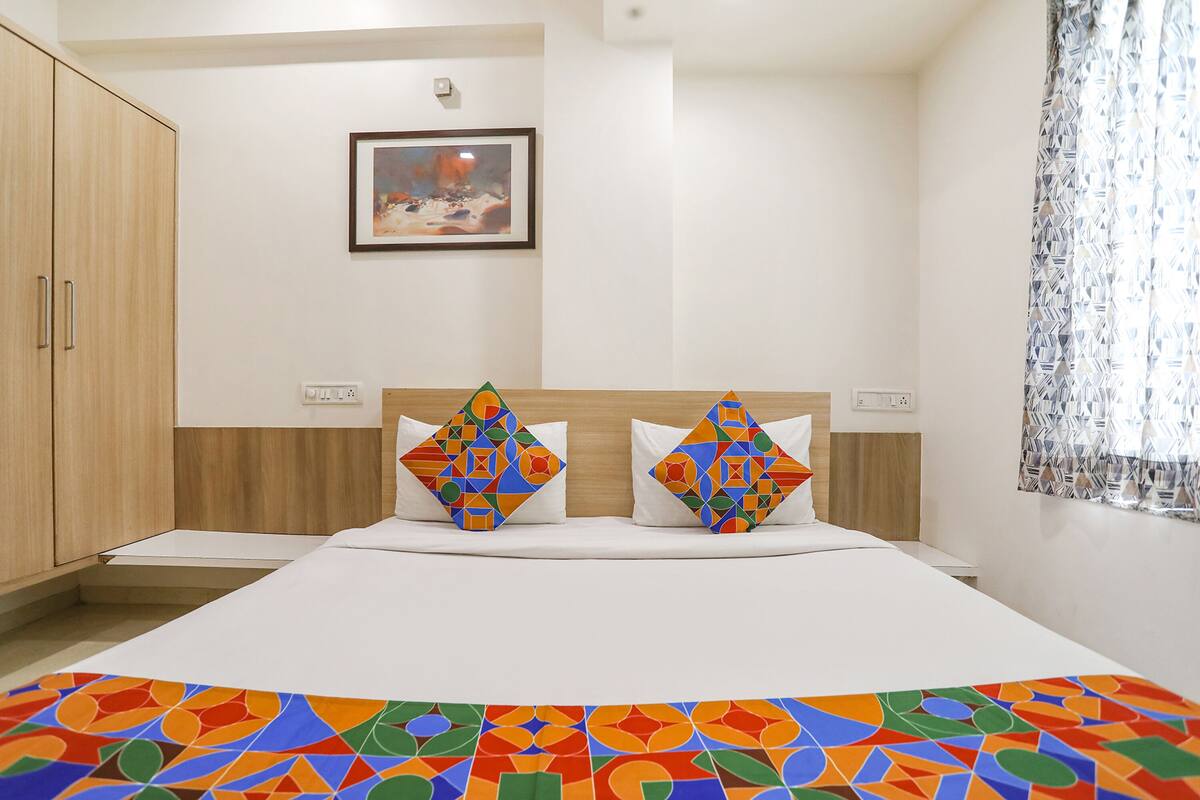 A hotel room with a bed, two pillows with geometric patterns, a framed picture on the wall, and a window with patterned curtains.