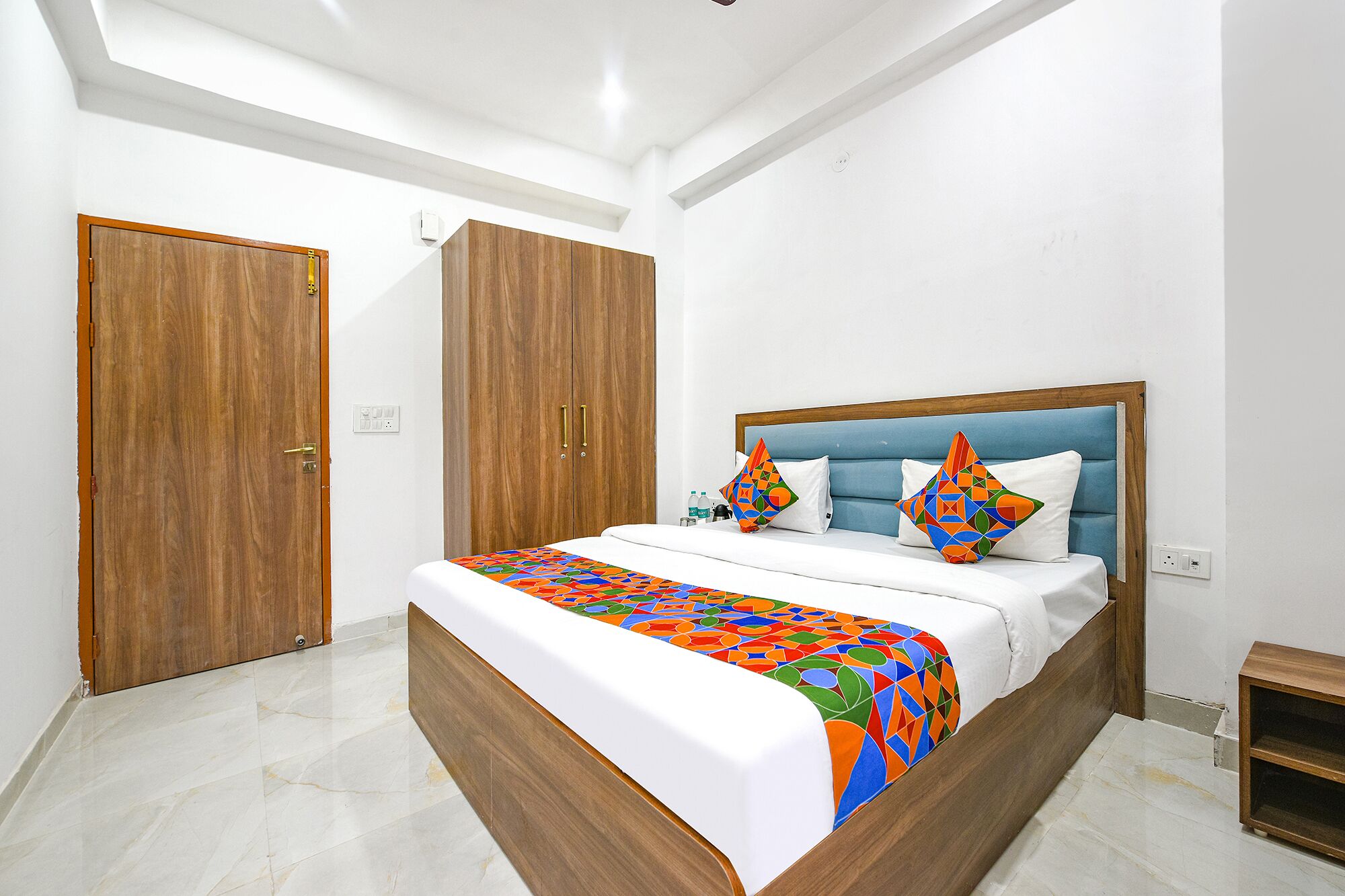 A hotel room with a bed, wooden wardrobe, and a small wooden shelf.