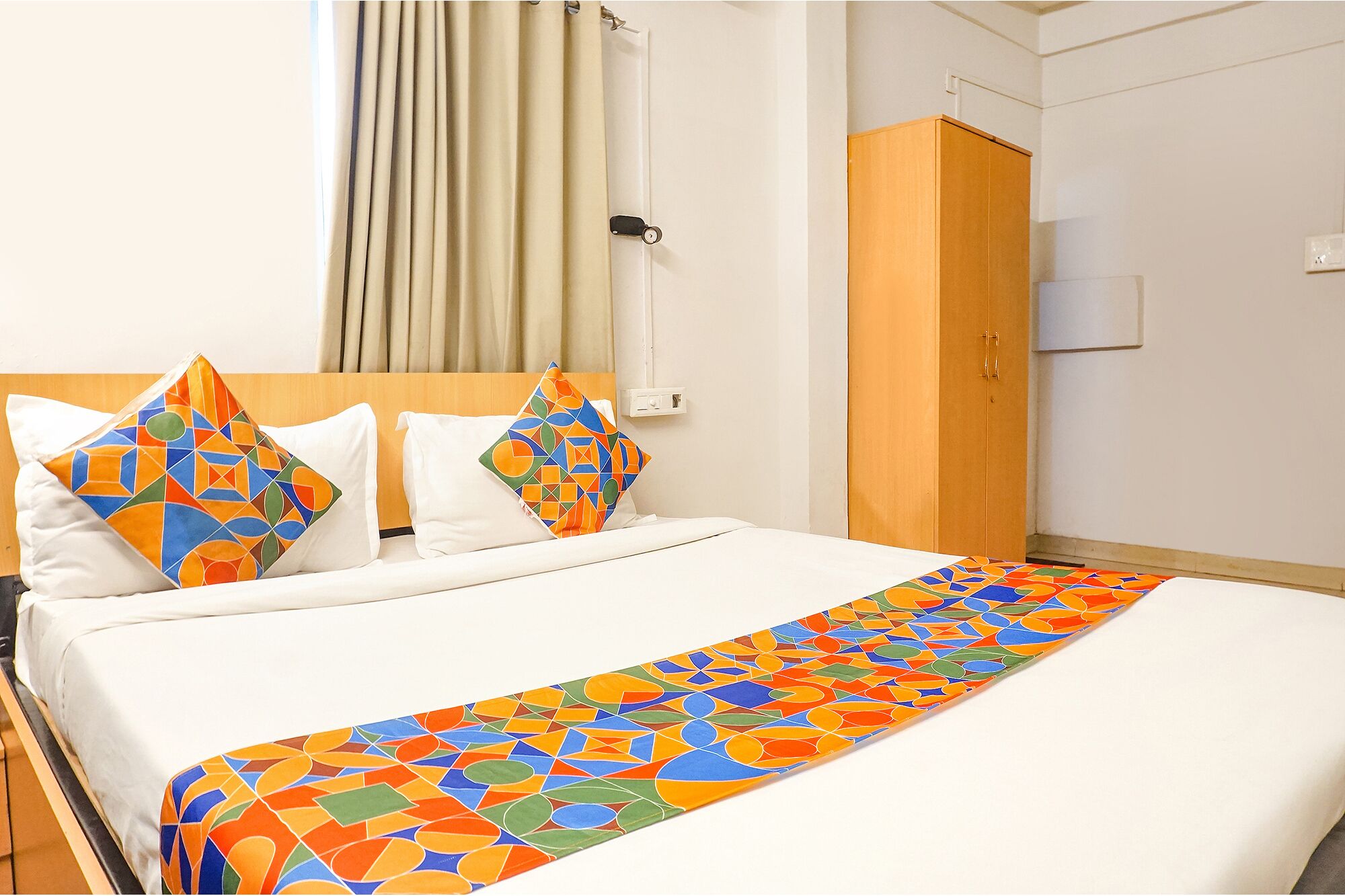A hotel room with a bed, two pillows with geometric patterns, a wooden wardrobe, and a window with curtains.